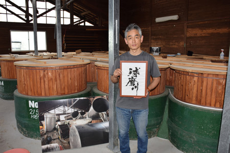 被災時に撮影した写真パネルと、「球磨川」を仕込んだタンクの前に立つ下田文仁さん=6月23日、熊本県人吉市